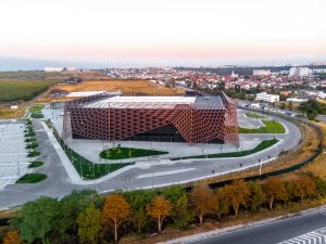 Aerial perspective of a large commercial building with a complex geometric roof design and surrounding roads and parking areas